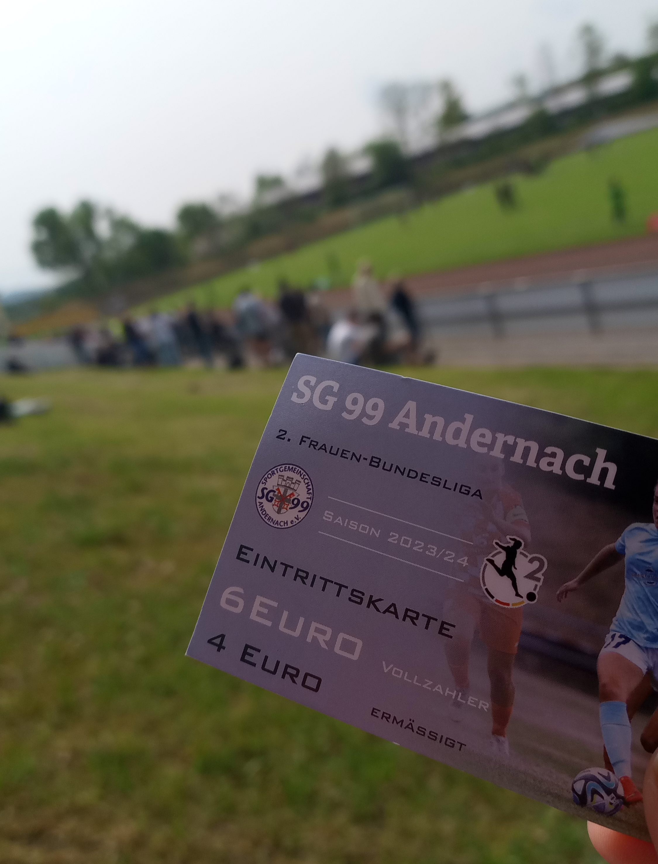 Im Vordergrund eine Eintrittskarte für das Heimspiel der SG Andernach Frauen gegen die Zweitvertretung der Eintracht. Im Hintergrund das Stadion Andernach. Viel Grün.
